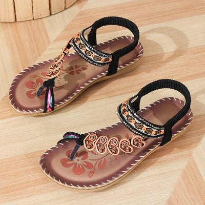 Sandales d'été pour femmes avec motifs floraux, semelle marron et lanières noires ornées de perles, posées sur un sol en bois clair.