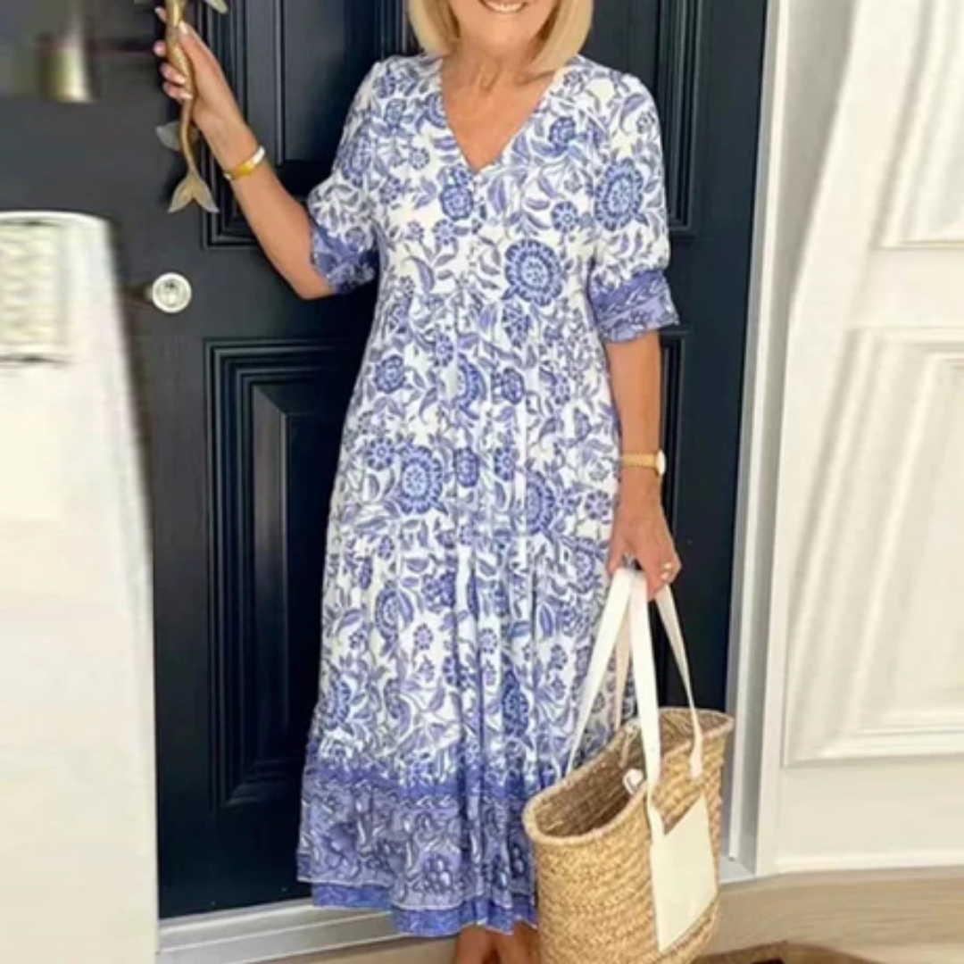 Robe longue bohème bleue et blanche à motifs floraux, femme tenant un sac en osier, devant une porte noire. Mode estivale élégante et décontractée.
