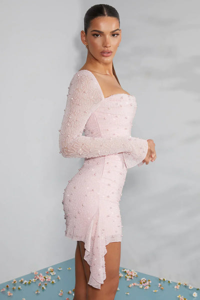 Robe rose pâle à manches longues ornée de perles, portée par une femme sur fond bleu. Mode élégante, tenue de soirée chic, style glamour.