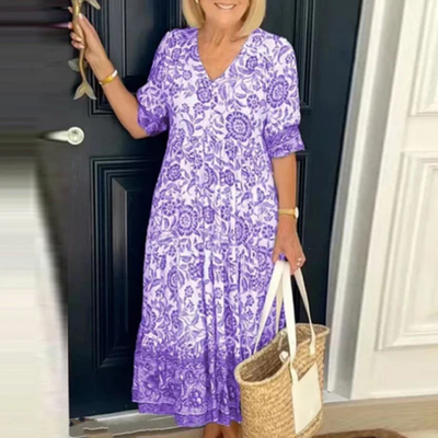 Femme en robe longue violette à motifs floraux, tenant un sac en osier, devant une porte noire. Mode estivale élégante et décontractée.