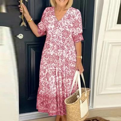 Femme en robe longue à motifs floraux roses, tenant un sac en osier, debout devant une porte noire. Mode estivale élégante et décontractée.