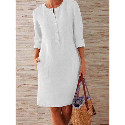 Robe blanche en lin pour femme avec manches longues retroussées, poches avant, et boutons, accompagnée d'un sac en osier. Mode estivale élégante.
