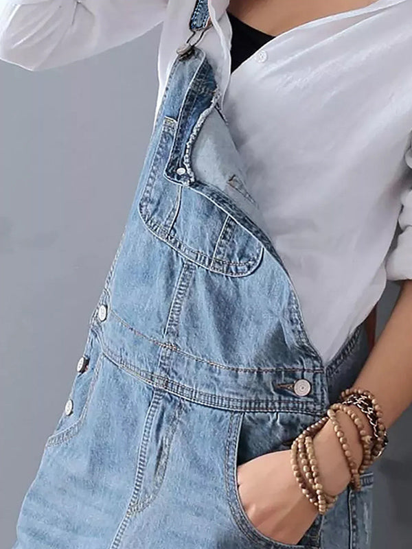 Personne portant une salopette en jean bleu avec une chemise blanche, main dans la poche, bracelets en bois au poignet. Mode décontractée tendance.