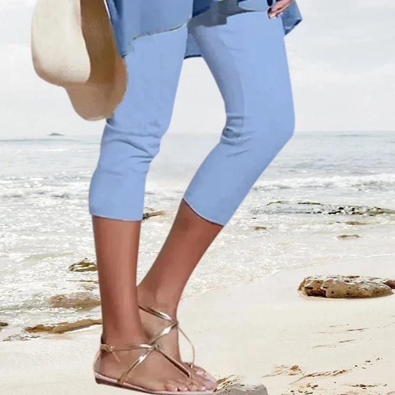 Femme en pantalon bleu et sandales dorées marchant sur une plage, tenant un chapeau de paille. Mode estivale, tenue décontractée bord de mer.