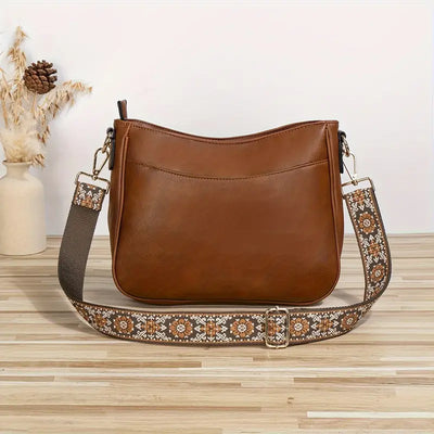 Sac à main en cuir marron avec bandoulière brodée florale, posé sur une table en bois clair, décor minimaliste avec vase et pommes de pin.