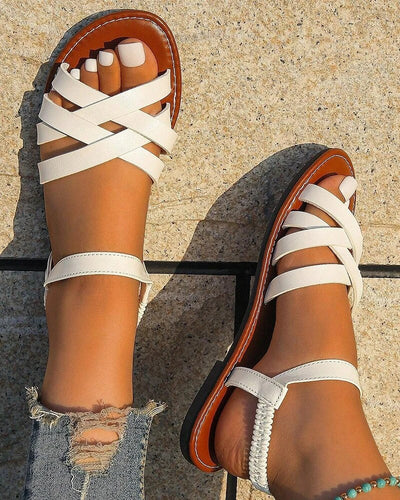 Sandales blanches en cuir croisées sur pieds féminins, posées sur un sol en pierre. Ongles des pieds vernis blanc, jean déchiré. Mode estivale élégante.