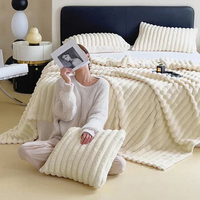 Femme en pull blanc assise sur un lit avec couverture et oreillers en peluche crème, tenant une photo. Décor moderne et minimaliste.