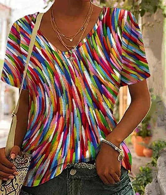 Femme portant un t-shirt coloré à motifs abstraits, col en V, manches courtes, avec un collier doré et un sac à main, en extérieur.