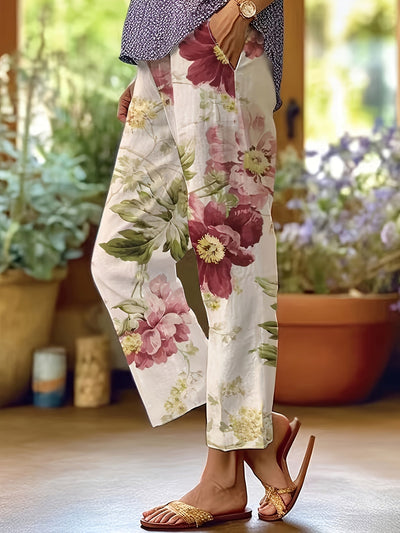 Pantalon femme à motifs floraux, fond blanc, fleurs rouges et vertes, style décontracté, sandales tressées, intérieur lumineux, plantes en pot.