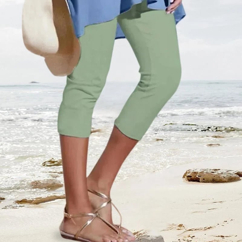 Femme en pantacourt vert et sandales dorées marchant sur une plage, tenant un chapeau de paille. Mode estivale, tenue décontractée bord de mer.