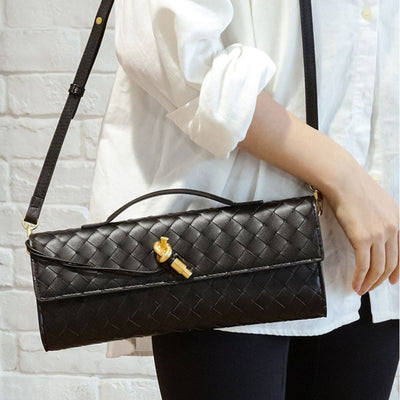 Sac à bandoulière noir en cuir tressé, porté par une personne en chemise blanche, avec une boucle dorée, sur fond de mur en briques blanches.