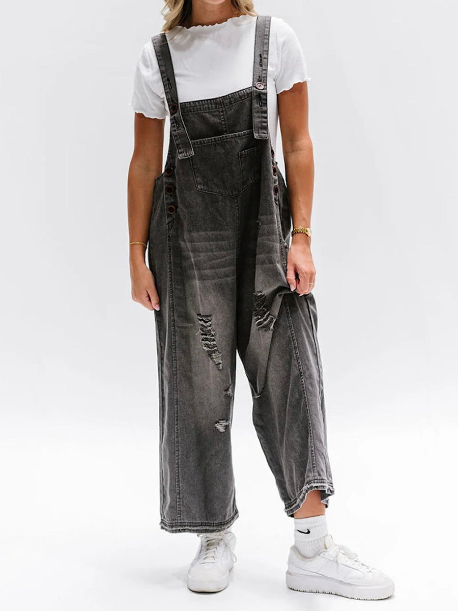 Salopette en jean gris délavé avec déchirures, portée par une personne en t-shirt blanc et baskets blanches, style décontracté et moderne.