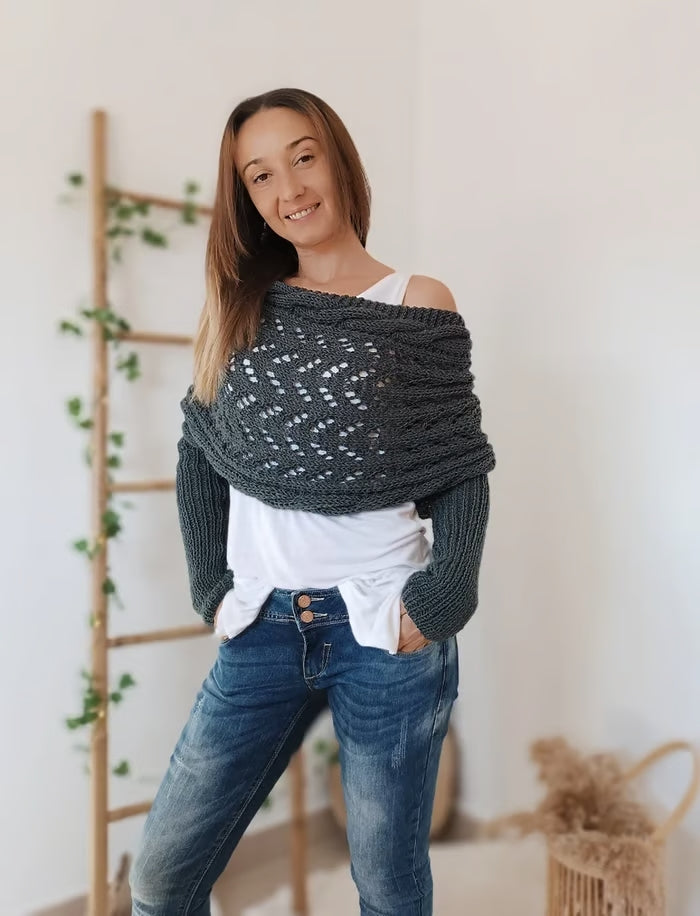 Jeune femme portant un pull tricoté gris avec motif ajouré, posant devant une échelle en bois décorée de plantes vertes. Mode automne-hiver tendance.