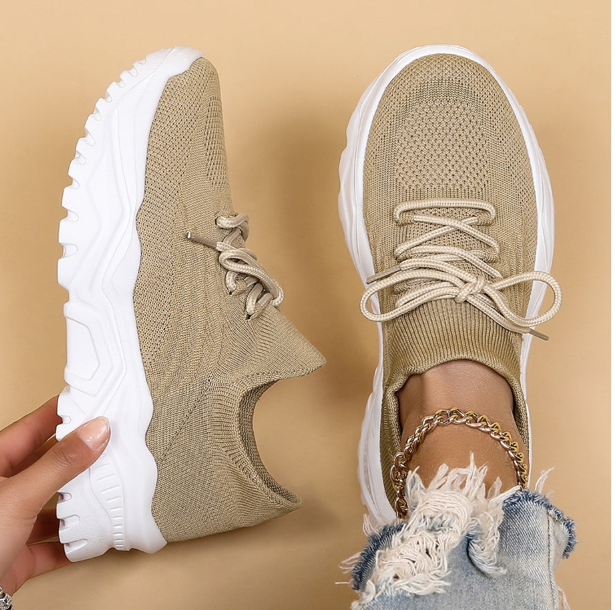 Chaussures de sport beiges en maille avec semelles blanches épaisses, tenues par une main, sur fond beige. Mode tendance, baskets confortables.