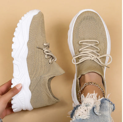 Chaussures de sport beiges en maille avec semelles blanches épaisses, tenues par une main, sur fond beige. Mode tendance, baskets confortables.