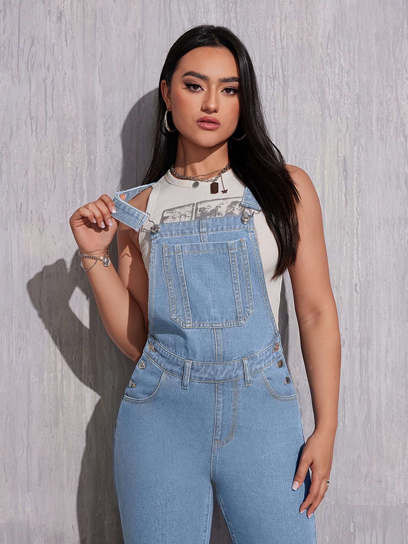 Femme portant une salopette en jean bleu clair, t-shirt blanc, posant devant un mur texturé. Mode décontractée, tendance denim, style urbain.