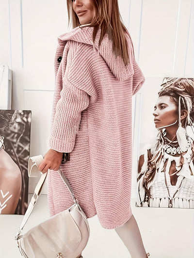 Femme portant un manteau rose en tricot, sac à main beige, posant devant des portraits en noir et blanc. Mode automne-hiver, style chic et moderne.