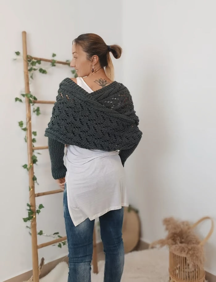 Femme portant un châle en tricot gris foncé, debout près d'une échelle en bois décorée de feuillage, vêtue d'un haut blanc et d'un jean.