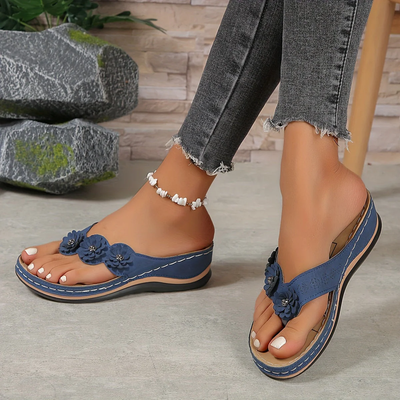 Sandales bleues à fleurs portées avec un jean gris effiloché, bracelet de cheville perlé, posées sur un sol en bois avec des pierres décoratives.