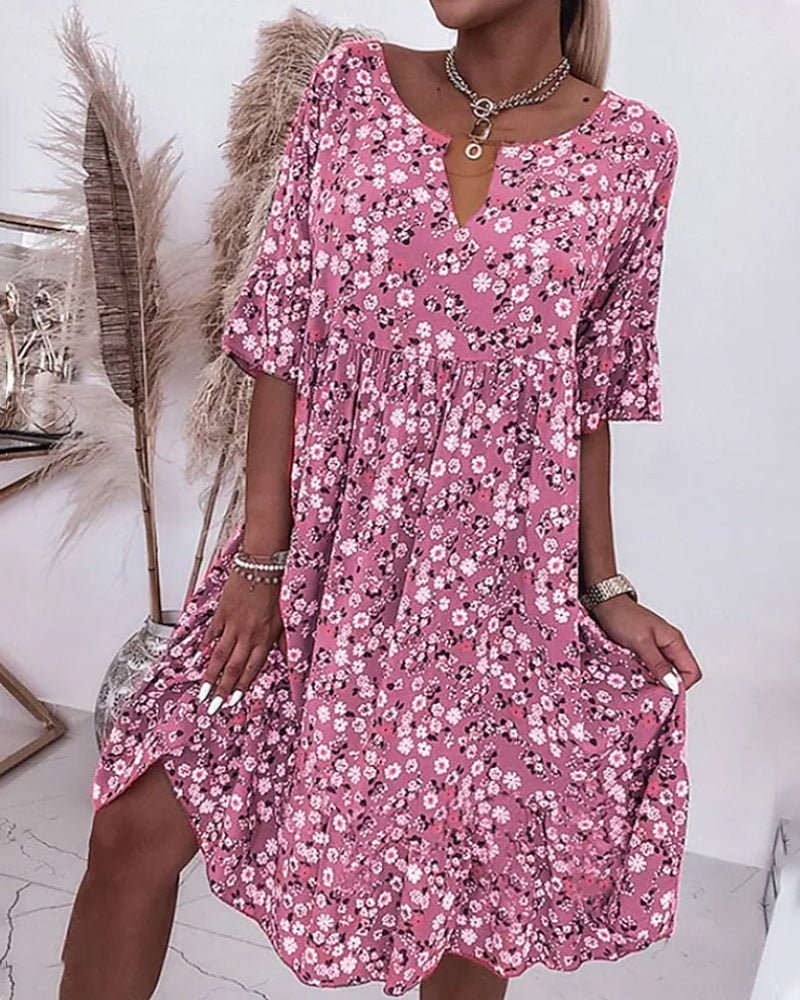 Robe rose à fleurs blanches et noires, manches mi-longues, col en V, portée par une femme. Décor avec plantes séchées en arrière-plan.