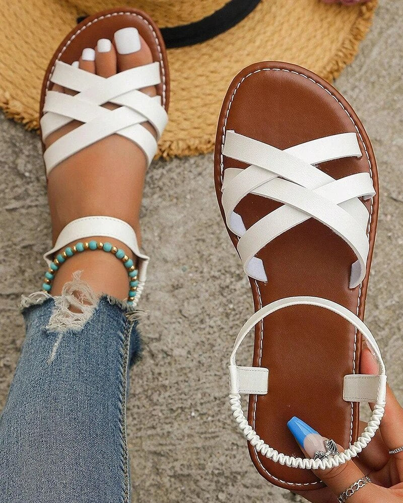 Sandales d'été blanches croisées sur pieds féminins, avec vernis à ongles blanc et bleu, bracelet turquoise, sur fond de chapeau de paille et jeans.