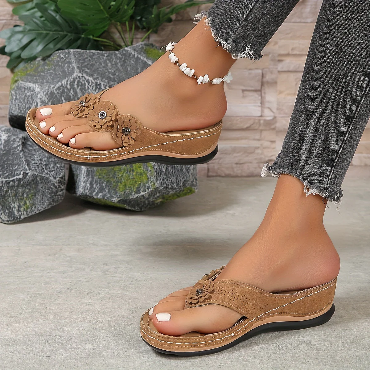 Sandales compensées marron avec fleurs décoratives portées par une femme, accompagnées d'un bracelet de cheville perlé, sur fond de pierres et plantes.