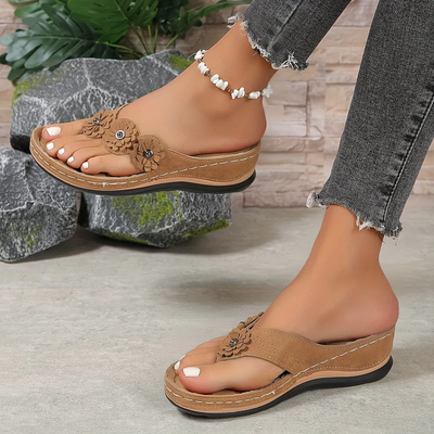 Sandales compensées marron avec fleurs décoratives portées par une femme, accompagnées d'un bracelet de cheville perlé, sur fond de pierres et plantes.