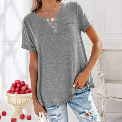 Femme portant un t-shirt gris à boutons, jeans déchirés, posant près d'un vase de pommes rouges dans un intérieur lumineux et élégant.
