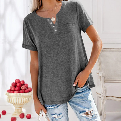 Femme portant un t-shirt gris à boutons, jeans déchirés, posant près d'un vase de pommes rouges. Mode décontractée, intérieur élégant.