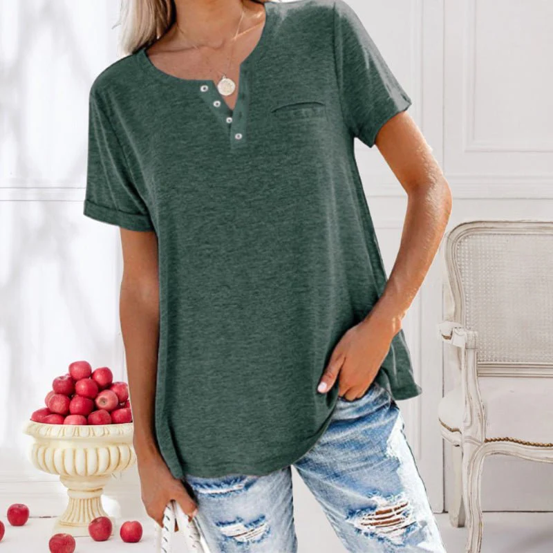 Femme portant un t-shirt vert à boutons, jeans déchirés, posant près d'un vase de pommes rouges. Mode décontractée, style estival, intérieur élégant.