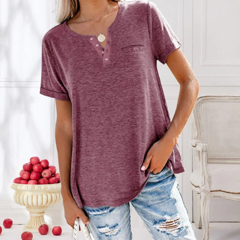 Femme portant un t-shirt bordeaux à col boutonné, posant dans un intérieur élégant avec des pommes rouges en arrière-plan. Mode décontractée chic.