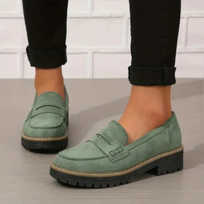 Chaussures mocassins vertes pour femme avec semelles épaisses noires, portées avec un pantalon noir. Style décontracté et moderne.
