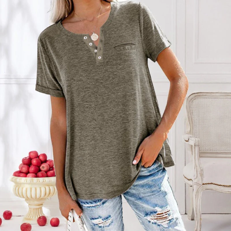 Femme portant un t-shirt gris à boutons, jeans déchirés, posant près d'un bol de pommes rouges dans un intérieur lumineux et élégant.