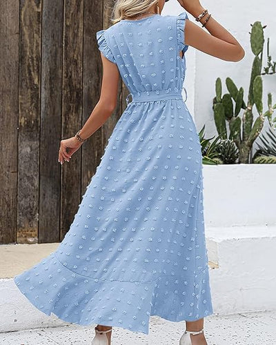 Robe longue bleue à pois en relief, manches à volants, portée par une femme de dos, devant un mur en bois et des cactus. Mode estivale élégante.