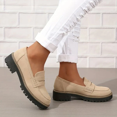 Chaussures mocassins beiges pour femme avec semelles épaisses noires, portées avec un jean blanc déchiré, sur fond de mur en briques blanches.