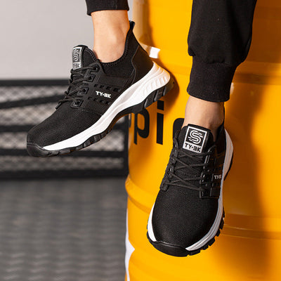 Chaussures de sport noires TY:3K pour homme, semelle blanche, portées sur un fond industriel jaune. Sneakers modernes et confortables.