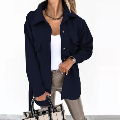 Femme portant une veste noire élégante, chemise blanche, pantalon beige, collier doré, tenant un sac à main à carreaux. Mode chic et moderne.