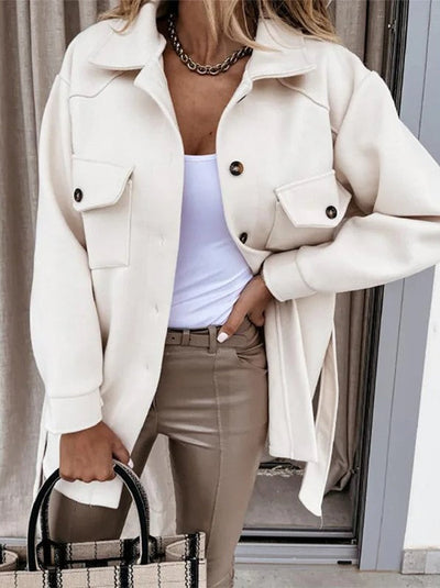 Femme portant une surchemise beige élégante avec boutons noirs, débardeur blanc, pantalon beige, collier chaîne, et sac à main rayé. Mode automne chic.