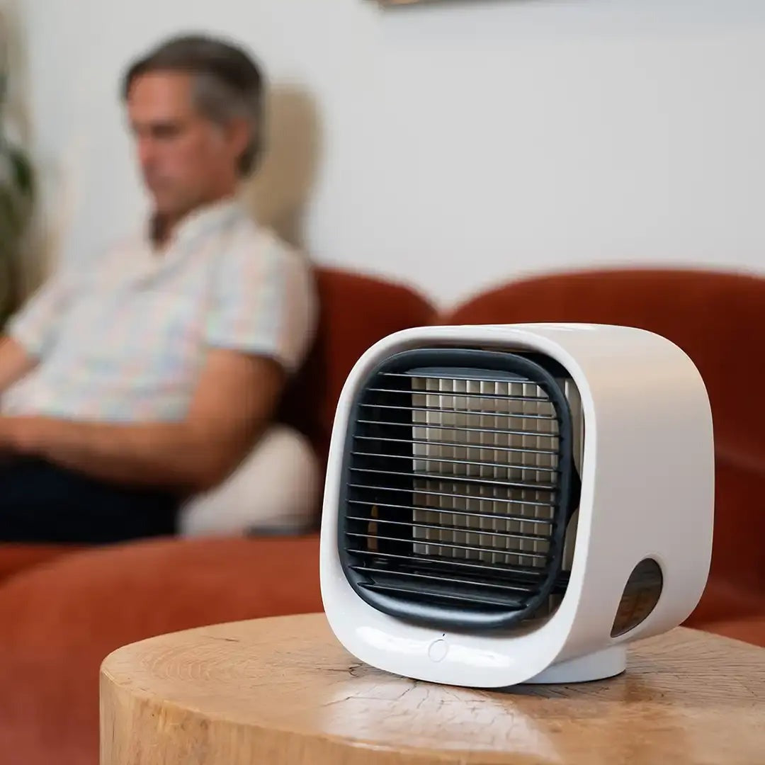 Homme assis sur un canapé flou en arrière-plan avec un mini ventilateur blanc moderne sur une table en bois au premier plan.