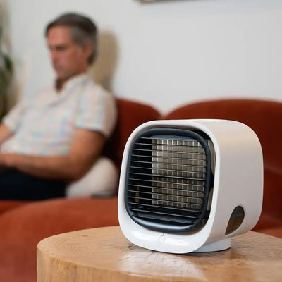 Homme assis sur un canapé flou en arrière-plan avec un mini ventilateur blanc moderne sur une table en bois au premier plan.