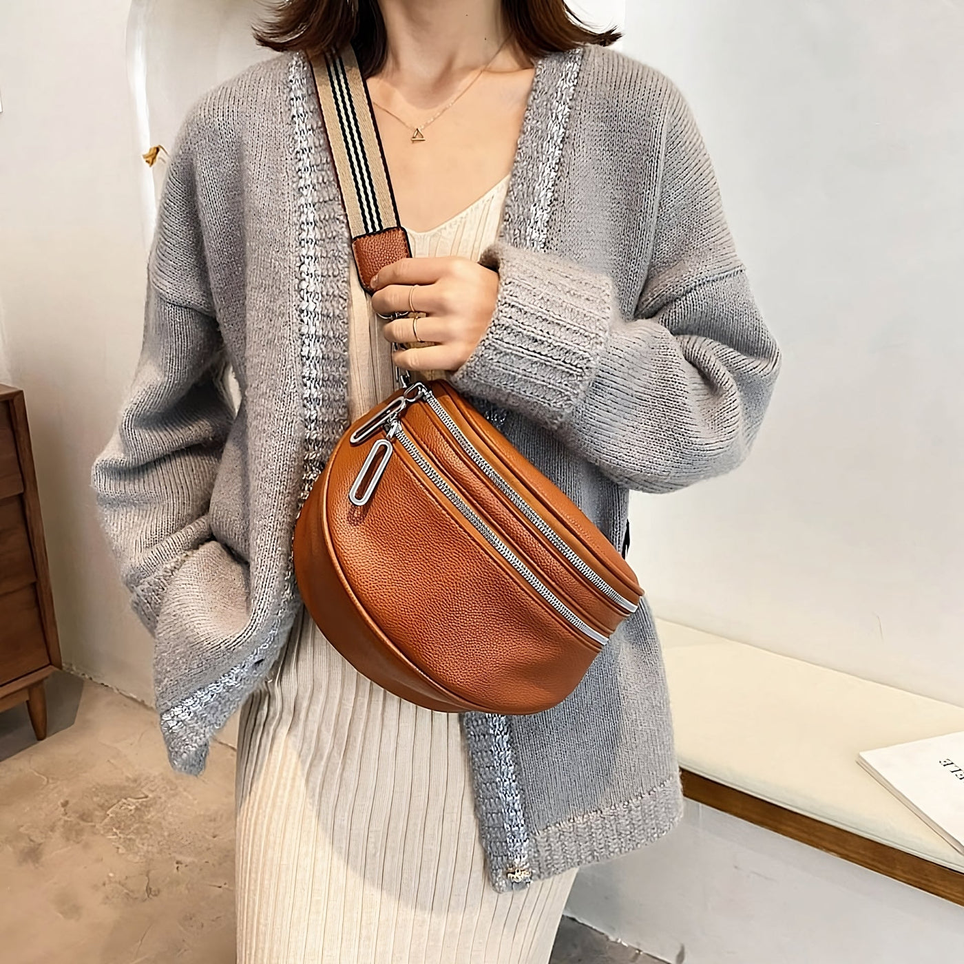 Femme portant un cardigan gris et une robe beige, tenant un sac banane en cuir marron avec bandoulière rayée. Mode automne, accessoires tendance.