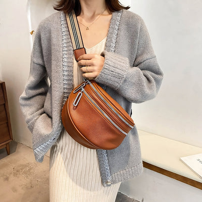 Femme portant un cardigan gris et une robe beige, tenant un sac banane en cuir marron avec bandoulière rayée. Mode automne, accessoires tendance.