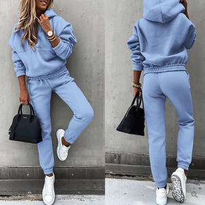 Femme en survêtement bleu clair, baskets blanches, sac à main noir, posant contre un mur gris. Mode décontractée, tendance streetwear.