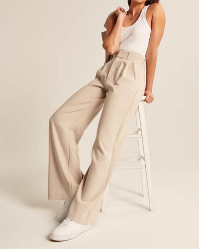 Femme en débardeur blanc et pantalon beige ample, posant sur un tabouret blanc, chaussures blanches; mode décontractée élégante.