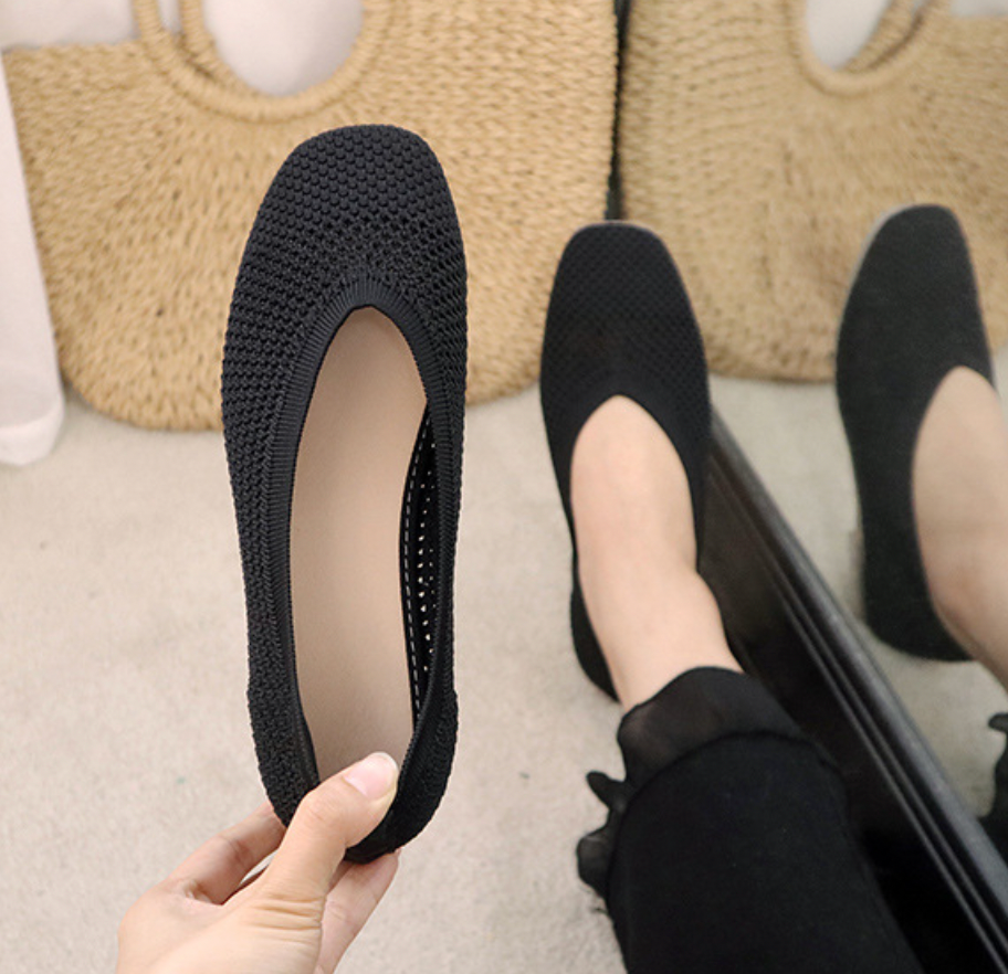Chaussures plates noires en tricot, confortables et élégantes, tenues par une main devant un miroir, avec un sac en paille en arrière-plan.