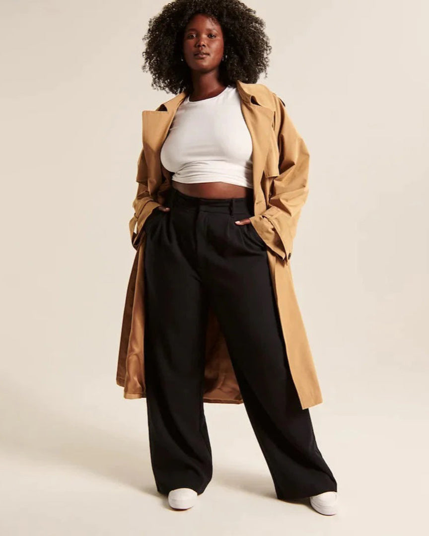 Femme portant un trench beige, un haut blanc et un pantalon noir ample, posant avec confiance. Mode automne, style décontracté chic.