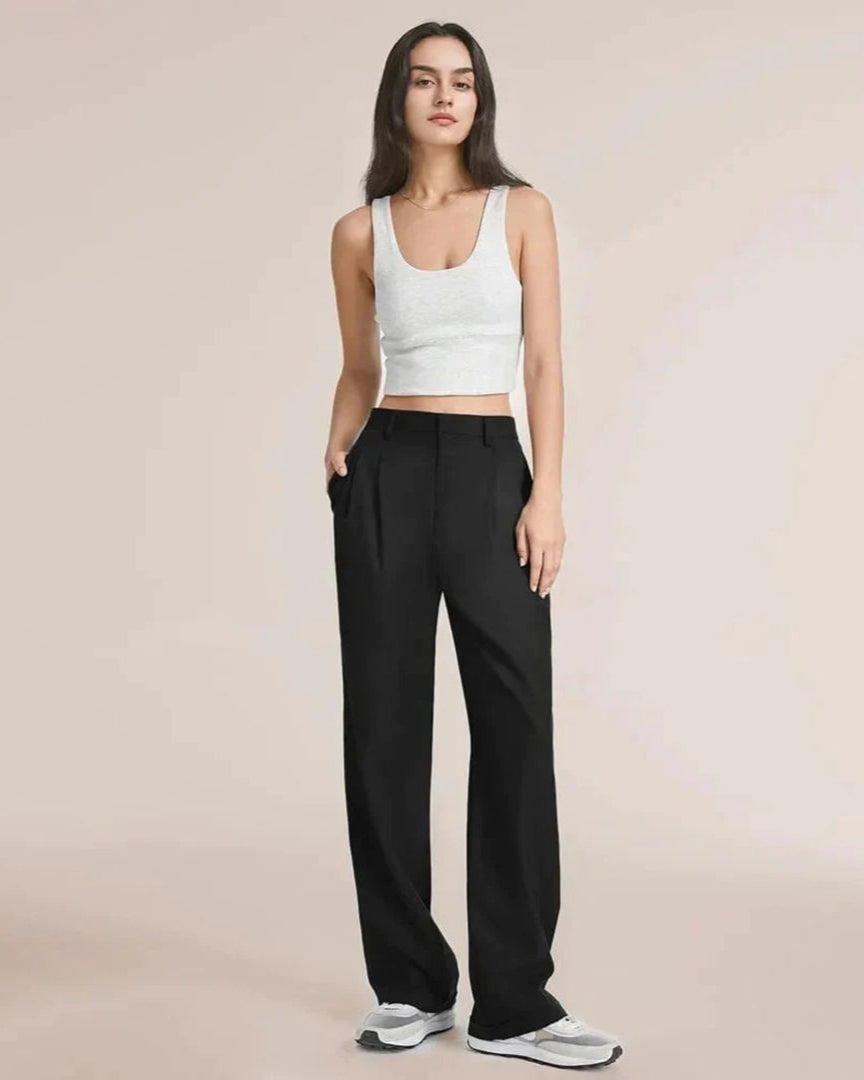 Femme portant un débardeur blanc et un pantalon noir ample, posant debout sur un fond neutre. Mode décontractée, élégance minimaliste.