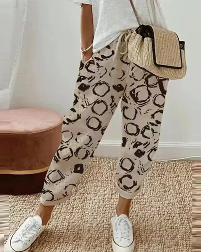 Femme portant un pantalon beige à motifs noirs, t-shirt blanc, sac en paille, baskets blanches. Mode décontractée, tendance, style urbain.