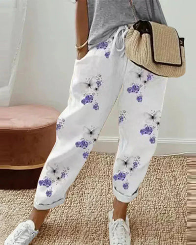 Pantalon blanc à motifs floraux violets, porté avec un t-shirt gris et un sac en paille. Style décontracté, mode féminine tendance.