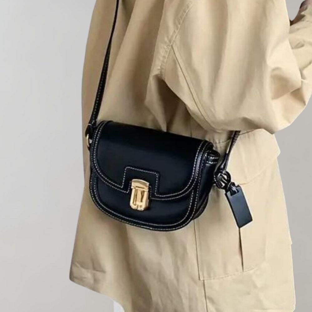 Sac à bandoulière noir en cuir avec fermoir doré, porté par une personne en manteau beige. Accessoire mode élégant et tendance.